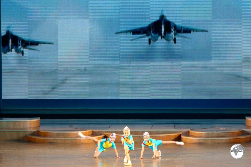 Girls performing a choreographed 'flight' routine in front of a screen displaying fighter jets.