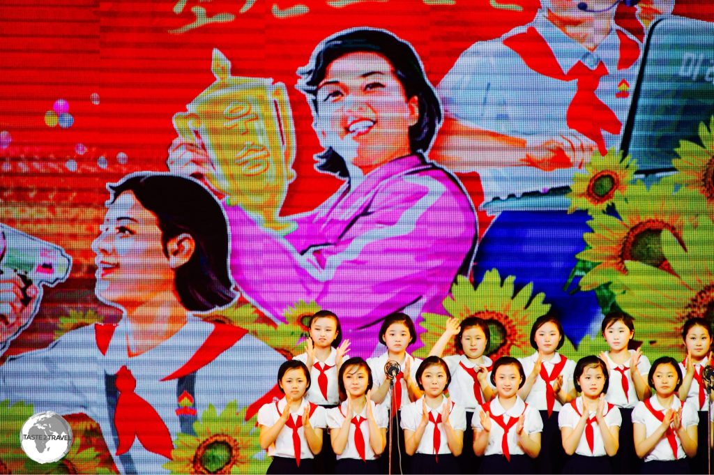 A girls choir performs in front of a propaganda poster.
