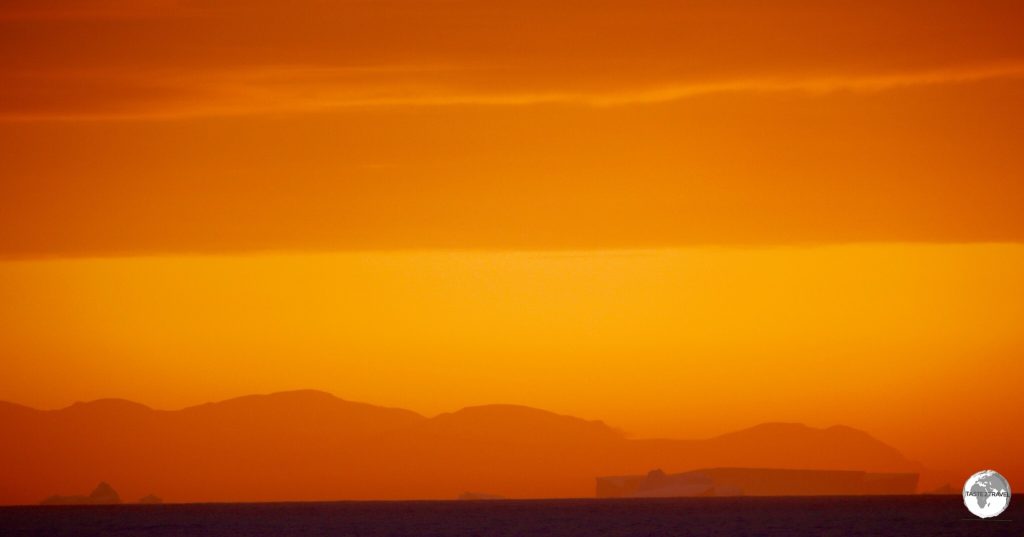 The rectangular shape of an iceberg stands out against a mountain range in the setting sun.