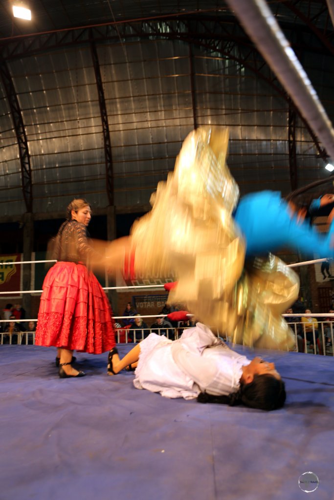 A golden swirl from the outer, plated skirt (la pollera) of an airborne Cholita, as she performs a body slam.